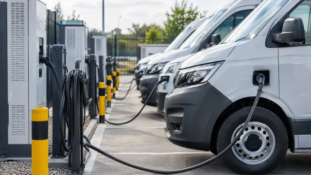 A row of electric delivery vans plugged into depot charge points, with charging cabinets and cable management posts behind them. AI-generated illustration.