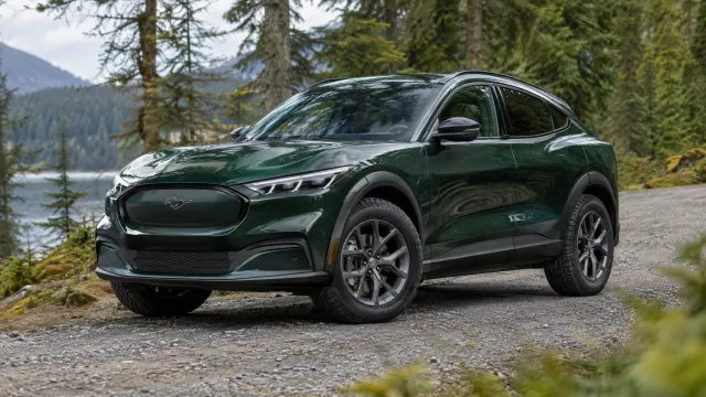 A 2027 Ford Mustang Mach-E Trail in dark green parked on a gravel forest road, showing its raised ride height, 19-inch wheels and Michelin CrossClimate 2 tyres. AI-generated illustration.