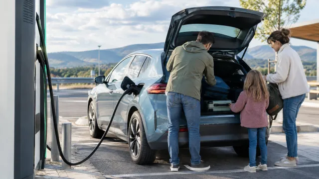 A compact electric hatchback plugged into a motorway fast charger in Europe, with a family unloading luggage from the boot. AI-generated illustration.
