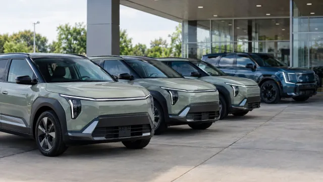A row of 2027 Kia EV3 crossovers parked outside a US dealership, with a three-row Kia EV9 visible behind them under the showroom canopy. AI-generated illustration.