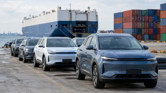 A row of used Chinese electric cars parked in a port holding area, with a car carrier and stacked shipping containers behind them. AI-generated illustration.